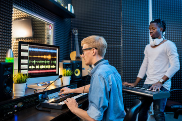 Young man recording music by computer while African guy playing pianoboard