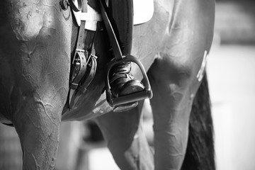 Black and white image of a rider's foot in the stirrup, which sits on a racehorse.