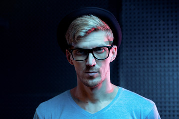 Serious young man in blue pullover, black hat and eyeglasses standing in studio