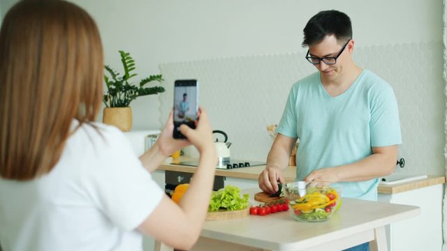 Joyful man husband is cooking salad cutting vegetables then juggling apples while girl recording video with smartphone camera at home in light kitchen.