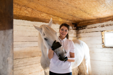 Happy young active woman embracing muzzle of purebred mare in stable