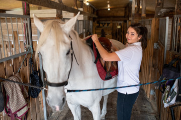 Young active woman putting saddle on back of white purebread racehorse