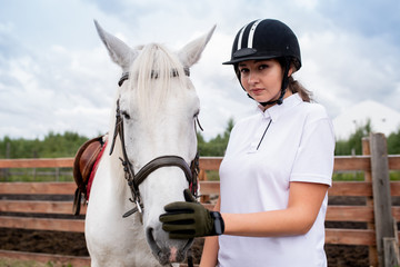 Pretty girl in equestrian helmet and gloves touching nose of white racehorse