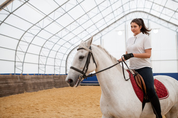 Serious young active woman looking straight while riding white purebred horse