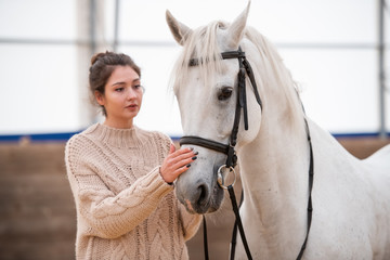 Young brunette woman in white knitted woolen sweater standing by white racehorse
