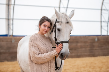 Pretty young brunette woman in casual sweater standing close to white horse