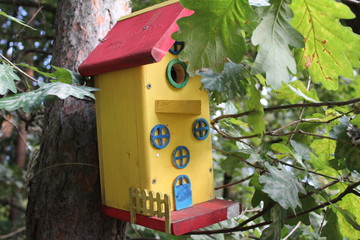 photo of the birdhouse for the birds in the Park.house made with your own hands.the material of wooden.the house is painted a yellow color.on it jewelry.time of year summer.trees with green foliage.