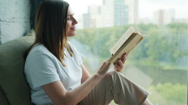 Slow motion of beautiful young woman reading book relaxing on window-sill at home enjoying leisure time in solitude. People, lifestyle and hobby concept.