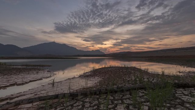 Timelapse dry land at the lake at Mengkuang Dam, Malaysia. A branch and dry land in the foreground. A hill and colorful sunset as background.