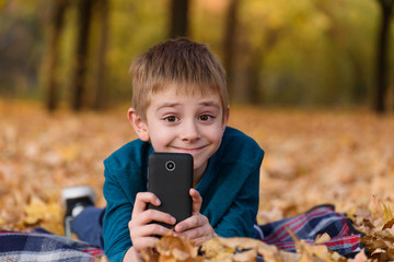 Cute schoolboy with smartphone lying on a plaid, yellow autumn leaves. Fall day