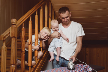 Father with a small child in his arms ironing linen. Eldest son is sitting nearby. Doing housework