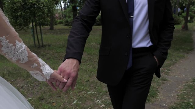 The bride and groom go by the hand, the bride in a wedding dress, a gold wedding ring, the groom in a black suit. Shooting with a 3-axis motorized gimbal stabilizer.
