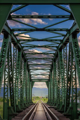 Vertical view through old, iron, railway bridge during sunset with dramatic, colorful clouds and sunlit other side