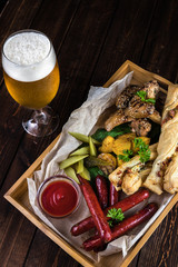 Oktoberfest food snacks concept top view. Wooden table with a glass of cold beer with water droplets and a big wooden box with grilled sausages, bread, pickles, fried chicken and potatoes