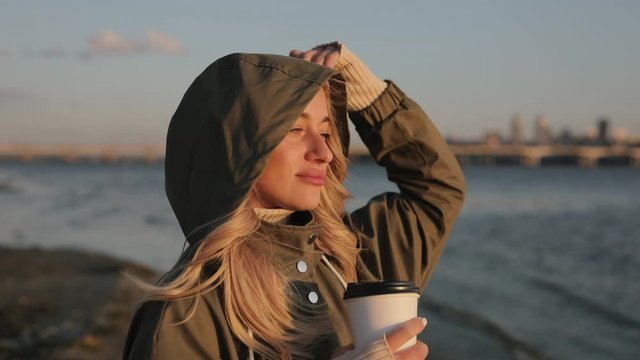 Woman in hoodie drinking tea standing near river enjoys autumn day