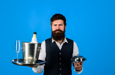 Waiter. Handsome waiter with serving tray and wine cooler. Waiter in restaurant carrying metal cloche lid cover and ice bucket with bottle, glass. Restaurant serving. Professional waiter in uniform.