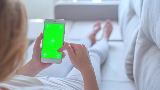 Hight quality 10bit footage of Young woman in white jeans uses SmartPhone with green screen laying on sofa at home. Made from 14bit RAW