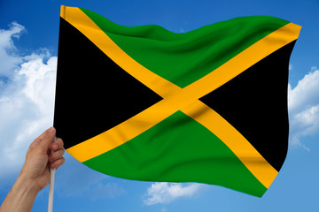 male hand holds against the background of the sky with clouds the national flag of Jamaica on a luxurious texture of satin, silk with waves, closeup, copy space, concept of travel, economy, politics