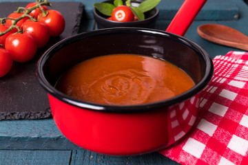 Molho de tomete na panela vermelha com fundo preto em cima da madeira preto e verde petróleo com colher de madeira, tomate cereja na ardósia negra e folhas de majericão