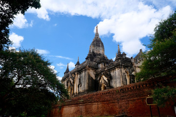 Looking up to magnificent ancient pagoda behind red brick wall. Under sunny blue sky white clouds. Green trees around. In Bagan Myanmar.