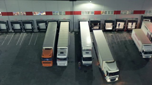 Aerial side view of a semi-trailers trucks standing at the ramps of the large logistics park with warehouse, loading hub for load/unload goods at night