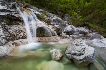 Alpine environment with a waterfall flowing between white rocks and into a emerald-green pond, surrounded by luxuriant green vegetation
