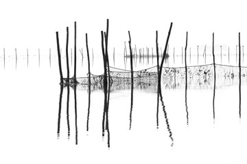 Black and white shot of a foggy lagoon with wooden poles and fishing nets reflecting on water
