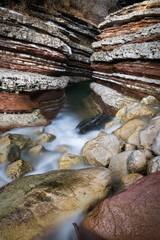 Entrance to a colorful and stratified slot canyon, with a small creek running in the middle; in foreground there are rocks and pebbles