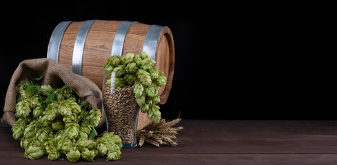 Barrel and beer glass with wheat and fresh green hops like a foam on dark wooden table. Empty space...