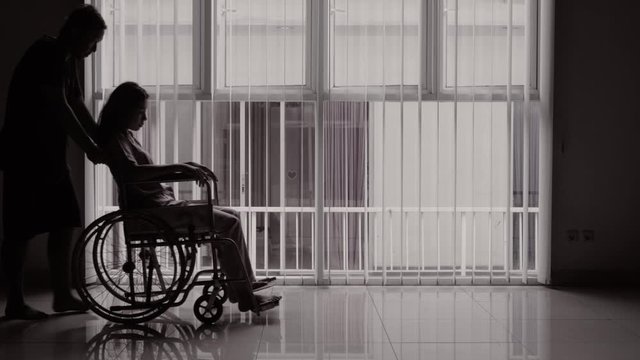 Silhouette of man helping his disabled wife by pushing the wheelchair at home. Shot in 4k resolution