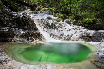 A small waterfall flowing into an emeral pond surrounded by white rock and green luxuriant vegetation © Roberto