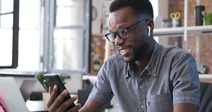 Happy african american man smiling and emotionally talking with his friend on smartphone. Man having video chat at office. Enjoying communicating. People and modern technology. Indoors 4k close up