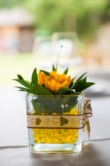 Close up of a tabletop decoration consisting in a square glass bowl containing a yellow sunflower against a bokeh background