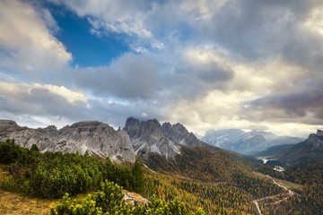 Alpine landscape with a valley covered with trees and distant mountains, under a blue sky with puffy clouds