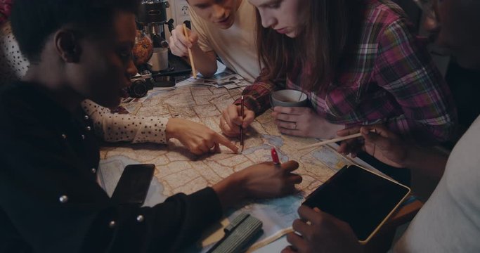 Close-up happy young multiethnic friends sit together looking at big map with pencils on kitchen table slow motion.