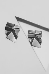 Closeup shot of metallic earrings that look like wrinkled squares made of silver metal. The pair of...