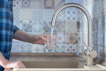 Woman in a blue plaid shirt pours water into the glass of the faucet in the kitchen