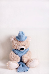 Children's toy teddy bear is sitting on a white sofa in a knitted scarf and hat in blue.