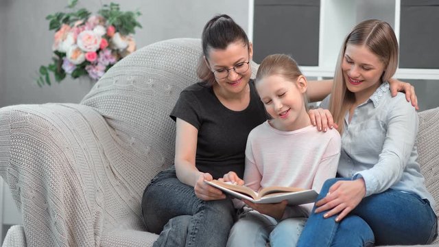 Two happy young smiling same sex mother reading book with their daughter at domestic interior