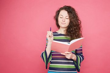 Beautiful curly haired young female office worker looks down, holds lilac pen, writes records, reminding note in notepad, makes planning on next week. Student prepares for college exam on pink wall.