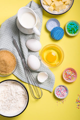 Baking ingredients for cake, cupcakes or muffins on yellow background, sugar pearls, eggs, whisk, flour, butter, sugar and milk