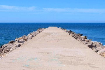 Fototapeta na wymiar Seascape background with empty long stone concrete pier stretching out to sea at sunny day. Cement pier with rocks on edges. Clear blue sky and water.