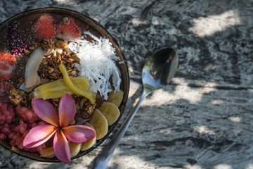 smoothie bowl in a coconut shell plate with with strawberries, chia seeds, banana, granola, flower on wooden background. Green tropical leaves background