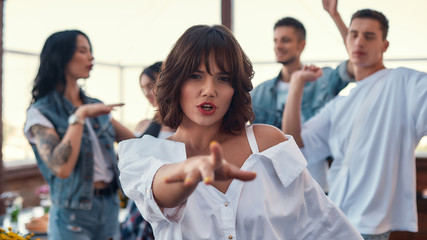 Obraz premium Dancing with friends. Pretty and young woman in white shirt is gesturing and looking at camera while standing on rooftop terrace with her friends