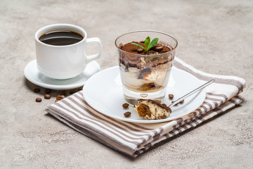 Portion of Classic tiramisu dessert in a glass and cup of coffee on concrete background
