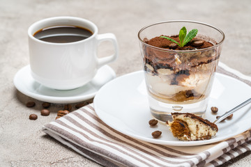 Portion of Classic tiramisu dessert in a glass and cup of coffee on concrete background