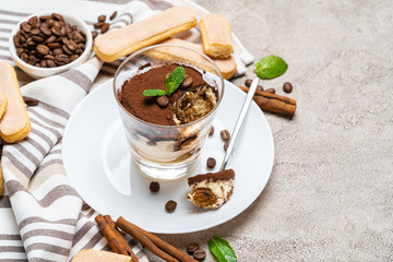 Portion of Classic tiramisu dessert in a glass, savoiardi cookies and cup of coffee on concrete background