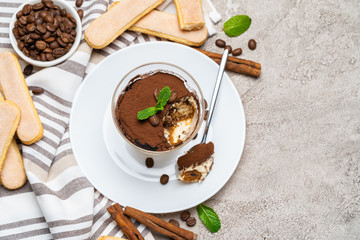 Portion of Classic tiramisu dessert in a glass, savoiardi cookies and cup of coffee on concrete background