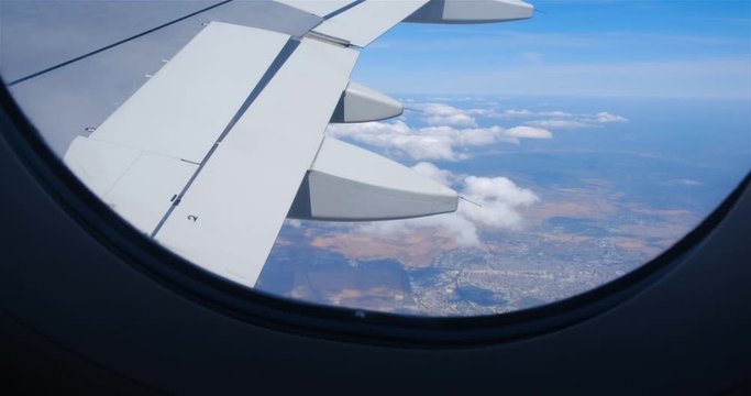 Close up of airplane right wing as seen from a passenger window seat while plane tilts down to reveal clouds and ground cities between brown crop fields in the Autumn.