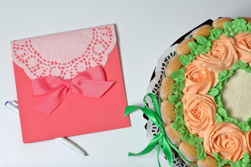 Sponge cake with cream cheese, decorated with Savoiardi cookies. On the side, wrapped in a ribbon tied to a bow. Near a greeting card and pen.